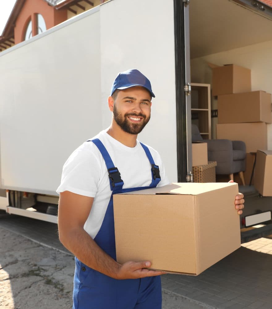 Smiling mover in a blue uniform carrying a cardboard box beside a loaded removal truck