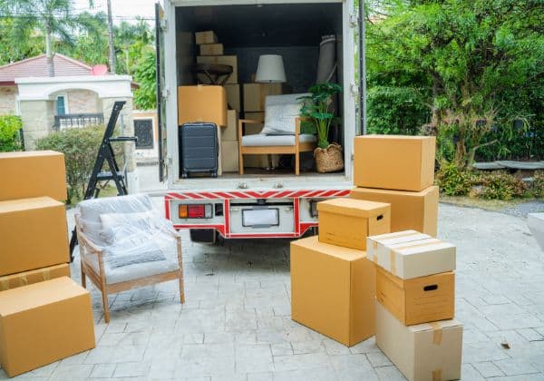 Open removal truck surrounded by stacked cardboard boxes and furniture on a driveway